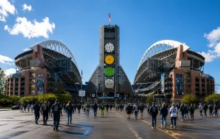 Seattle Seahawks fans walk toward Lumen Field on a sunny game day, with the stadium entrance and crowd visible under a bright blue sky.
