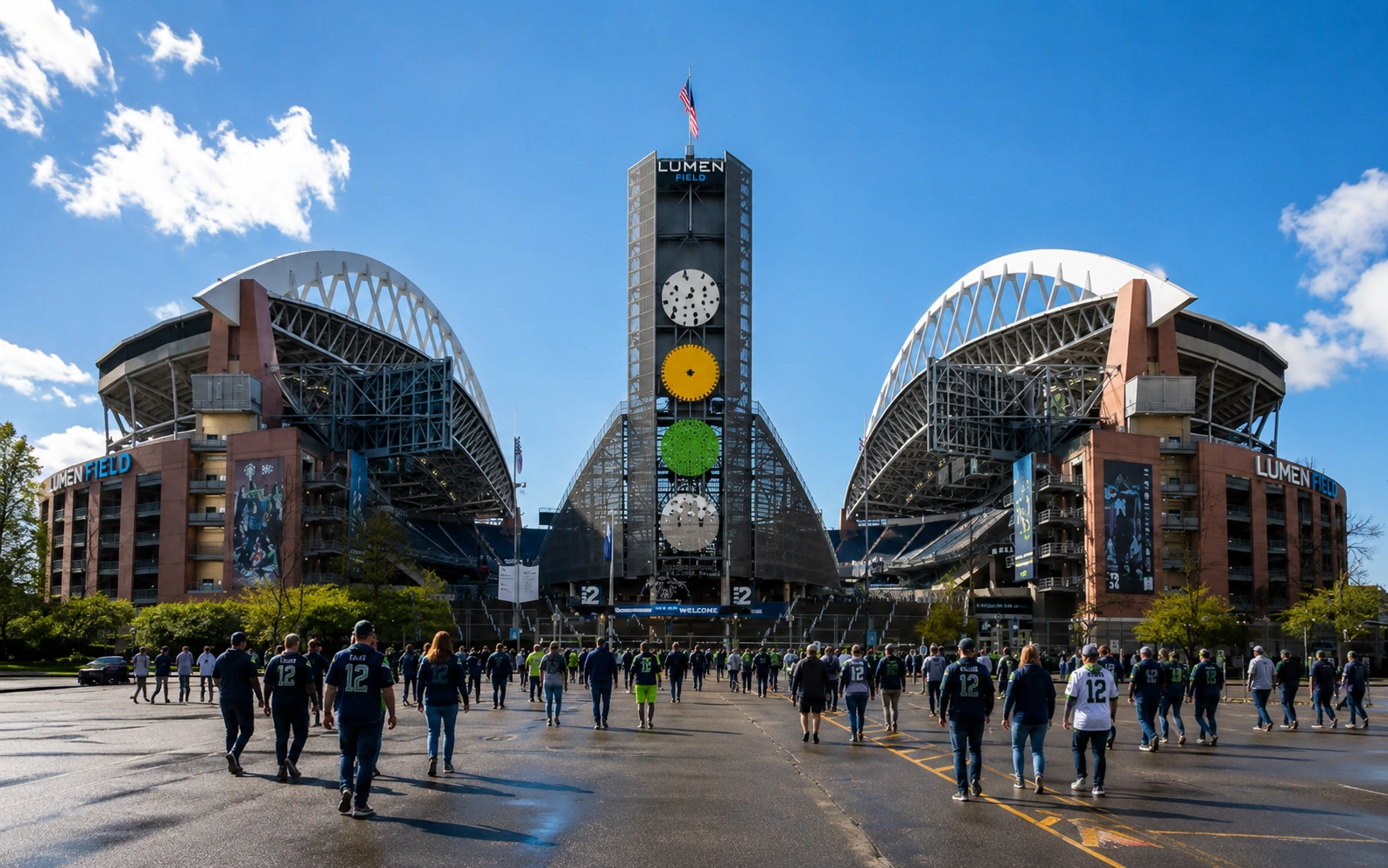 Seattle Seahawks fans walk toward Lumen Field on a sunny game day, with the stadium entrance and crowd visible under a bright blue sky.